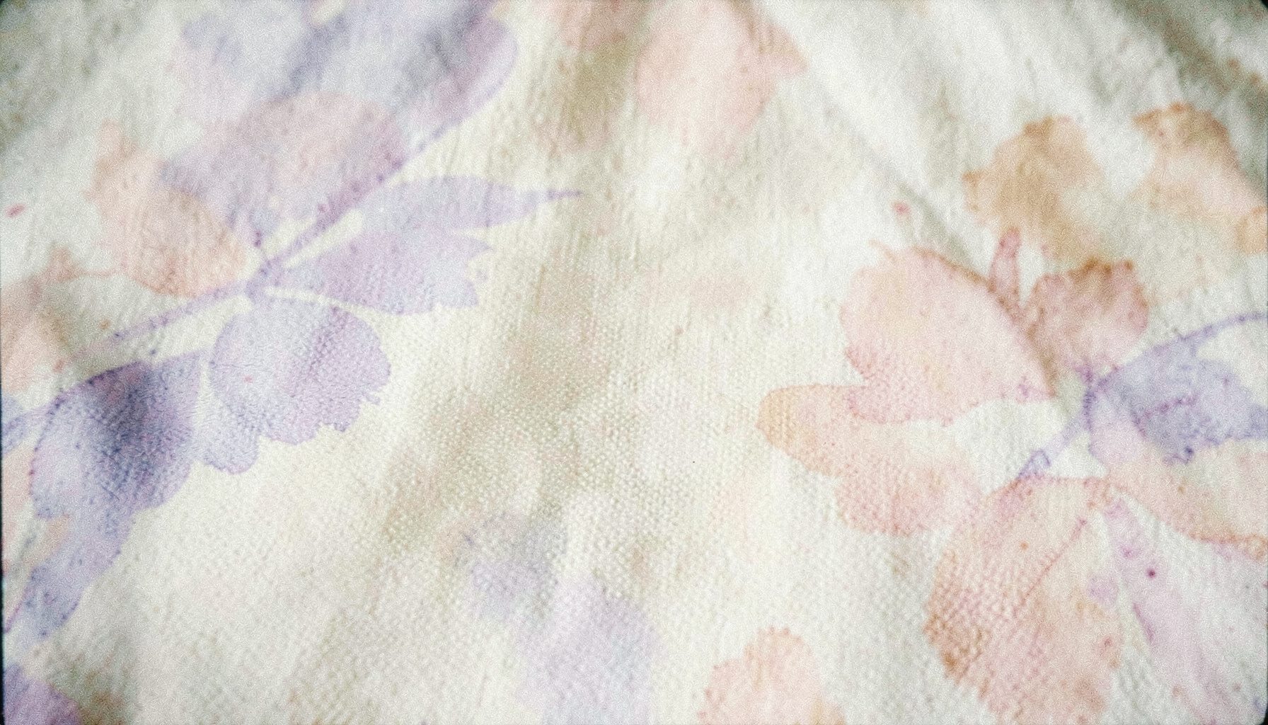 A softly lit analog film photograph with visible grain showing a textured cream fabric surface with subtle lavender and blush pink floral watercolor washes blending across the frame, calm and airy mood.
