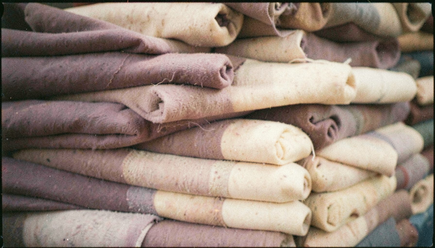 Analog film image with visible grain of neatly stacked textile rolls in muted dusty purple and cream tones, warm natural light.