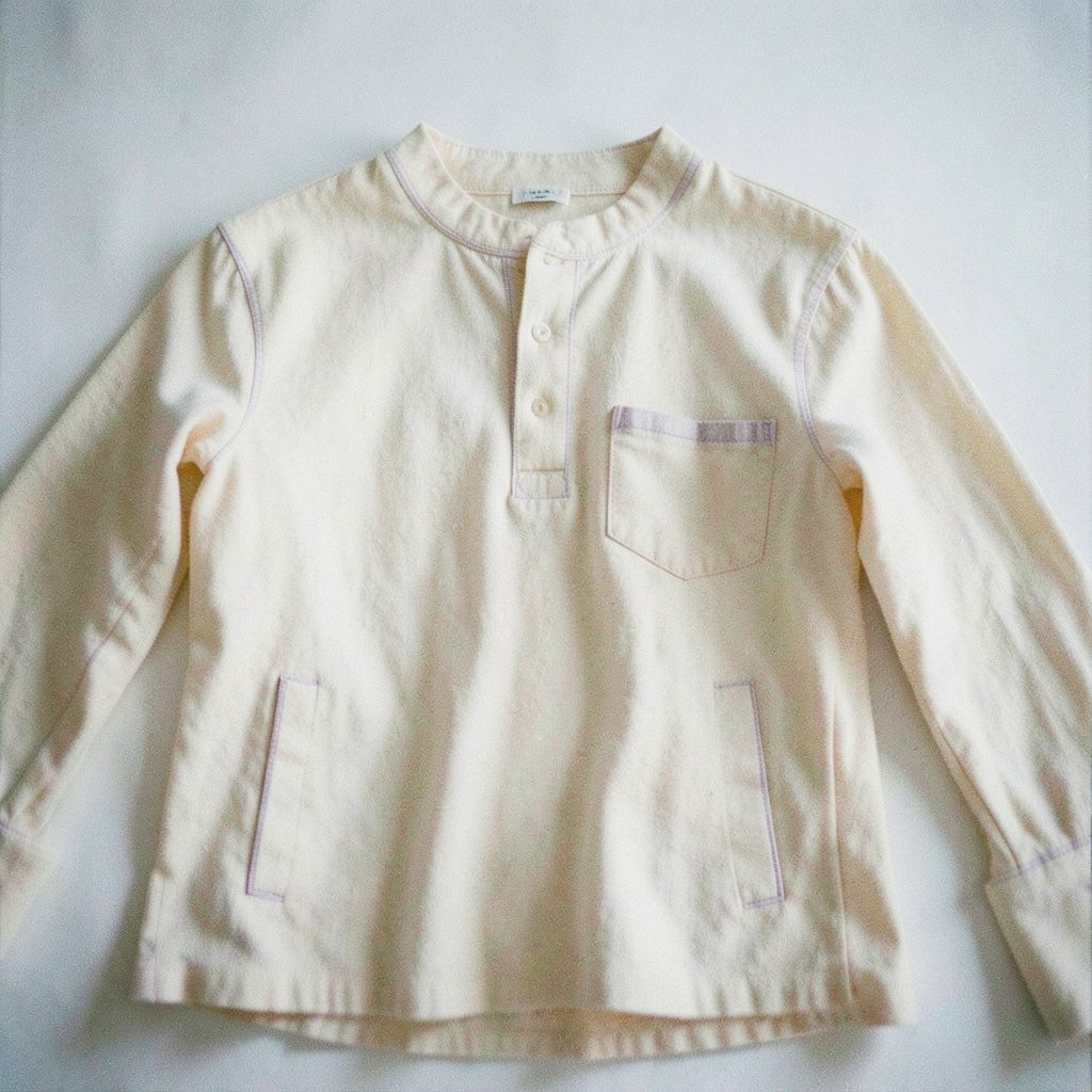 Film photograph with visible grain of a relaxed-fit garment in cream with subtle lavender stitching details, soft daylight.