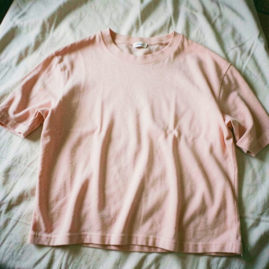 Grainy film photo of an oversized casual top in blush pink, draped naturally on a linen surface, airy and relaxed.
