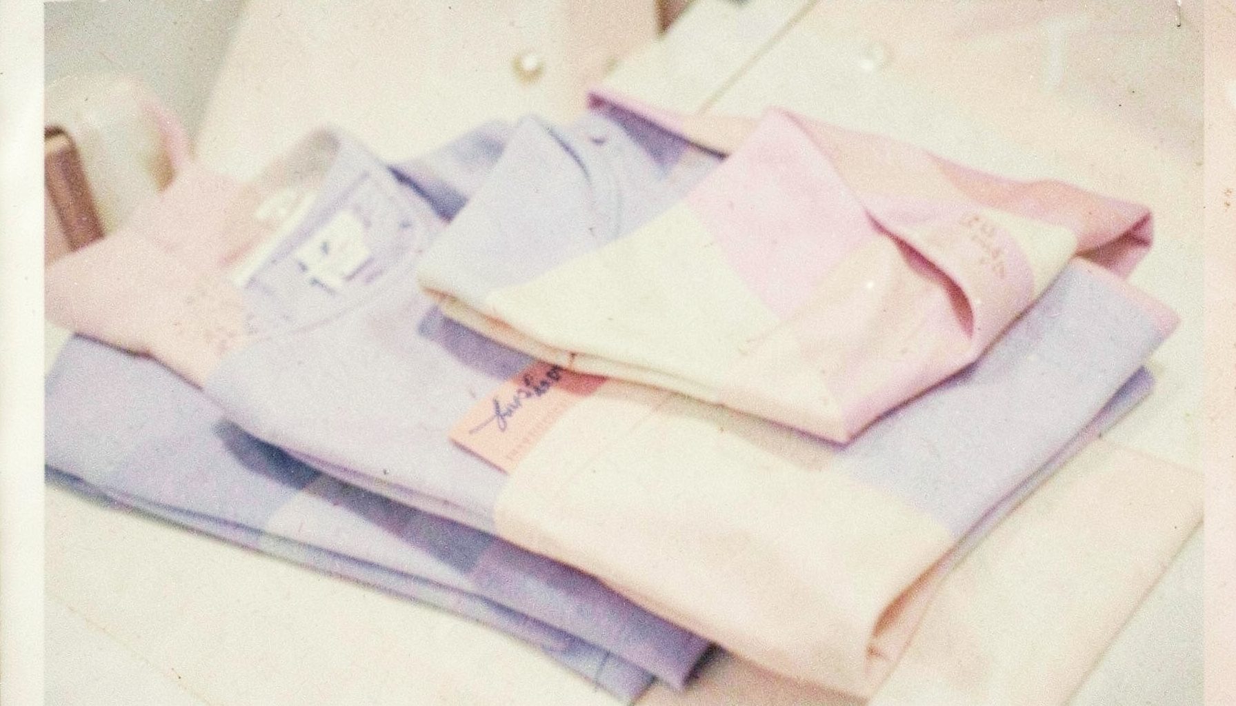 Grainy film photo of folded pastel-toned garments on a cream background, soft lavender and blush accents, gentle boutique atmosphere.
