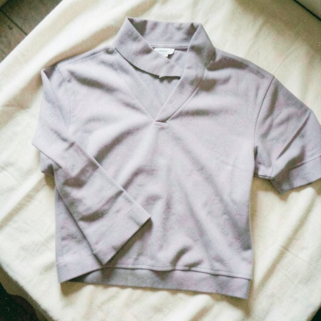 Grainy film photograph of a single folded jersey garment in soft lavender-gray on a cream linen surface, natural window light.