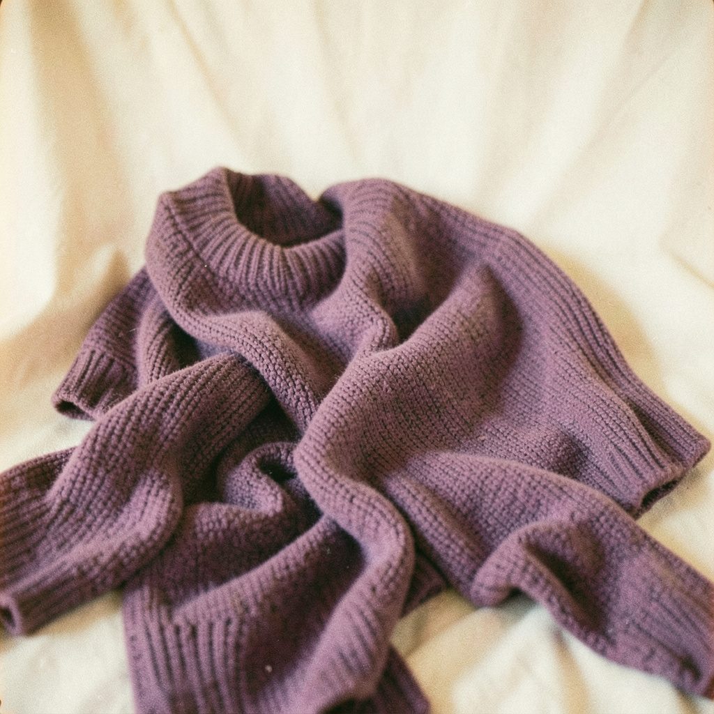 Soft film photo of textured knitwear in dusty purple hues, draped over a cream backdrop, warm and inviting mood.