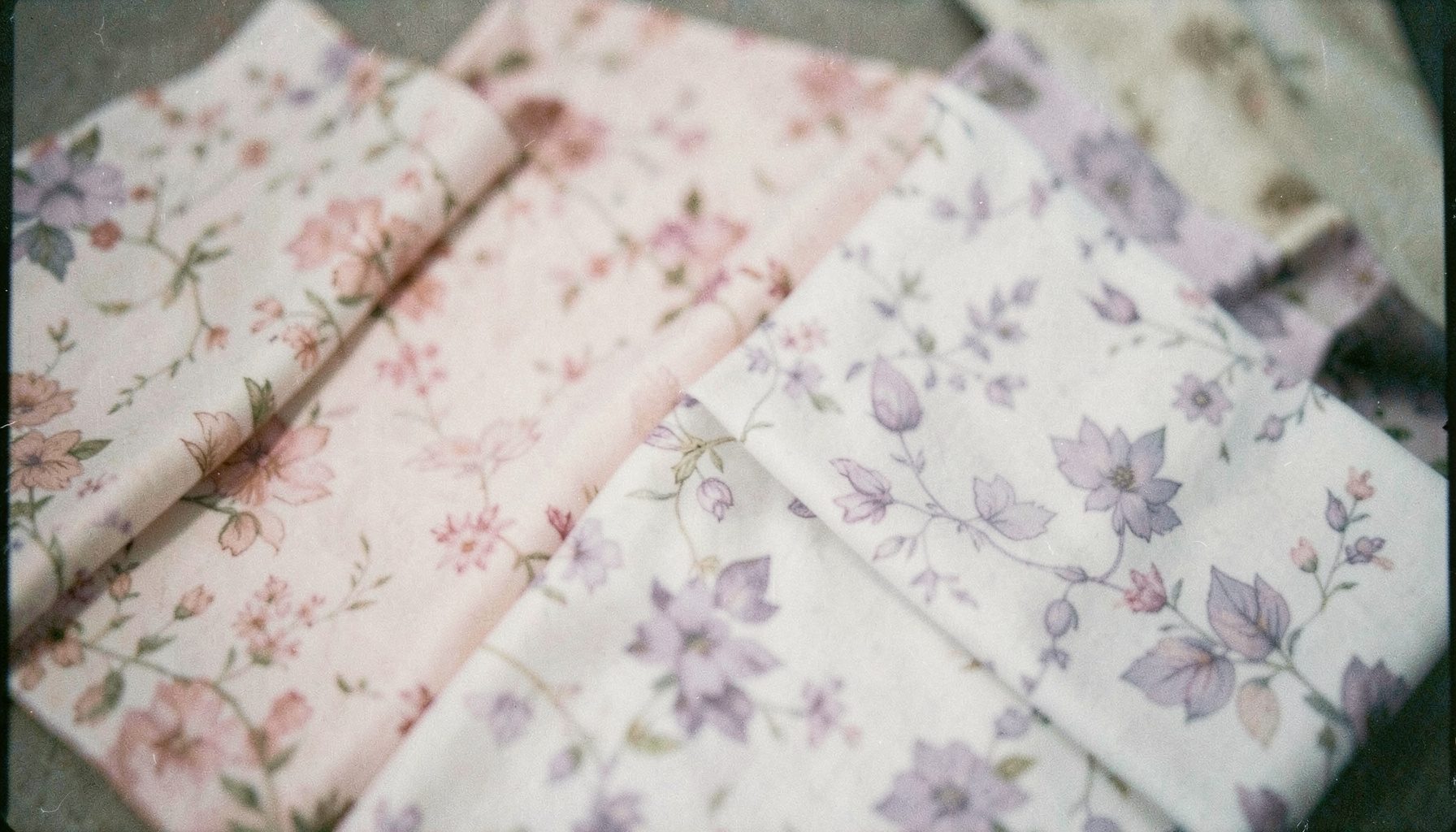 Soft-focus film photograph of delicate floral-patterned fabric swatches in blush pink and lavender, editorial boutique styling.