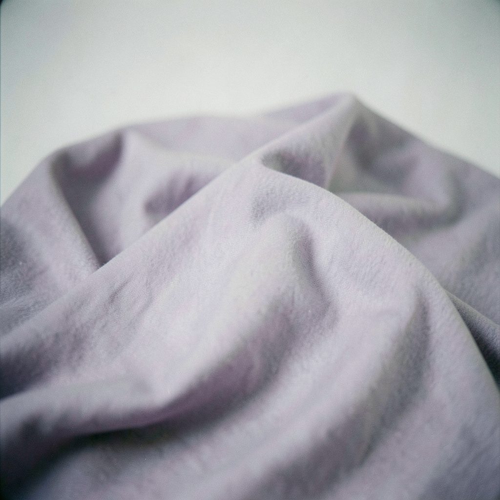 Soft-focus film photograph of performance-style fabric in lavender-gray, subtle texture visible, clean studio lighting.